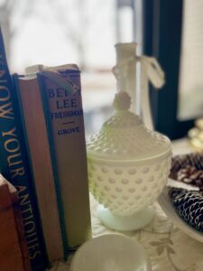 White Fenton hobnail milk glass glassware lidded dish styled with greenery and books on a vintage-inspired credenza.