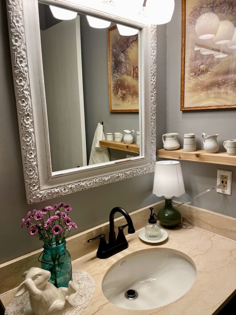 Vintage bathroom styled with thrifted decor including mirrors, art, and ironstone